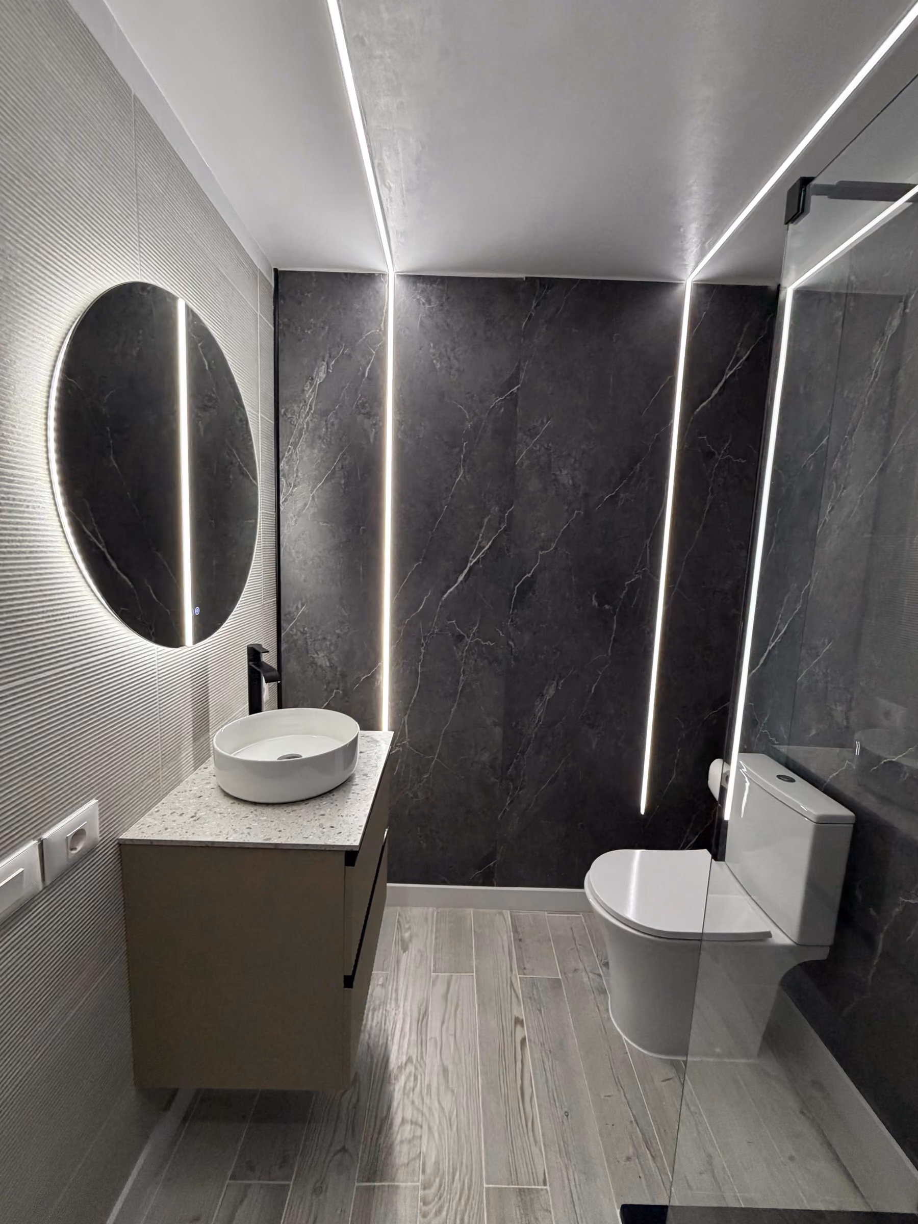 Baño en piedra oscura con ducha, espejo circular de marco negro y lavabo sobre encimera de terrazo