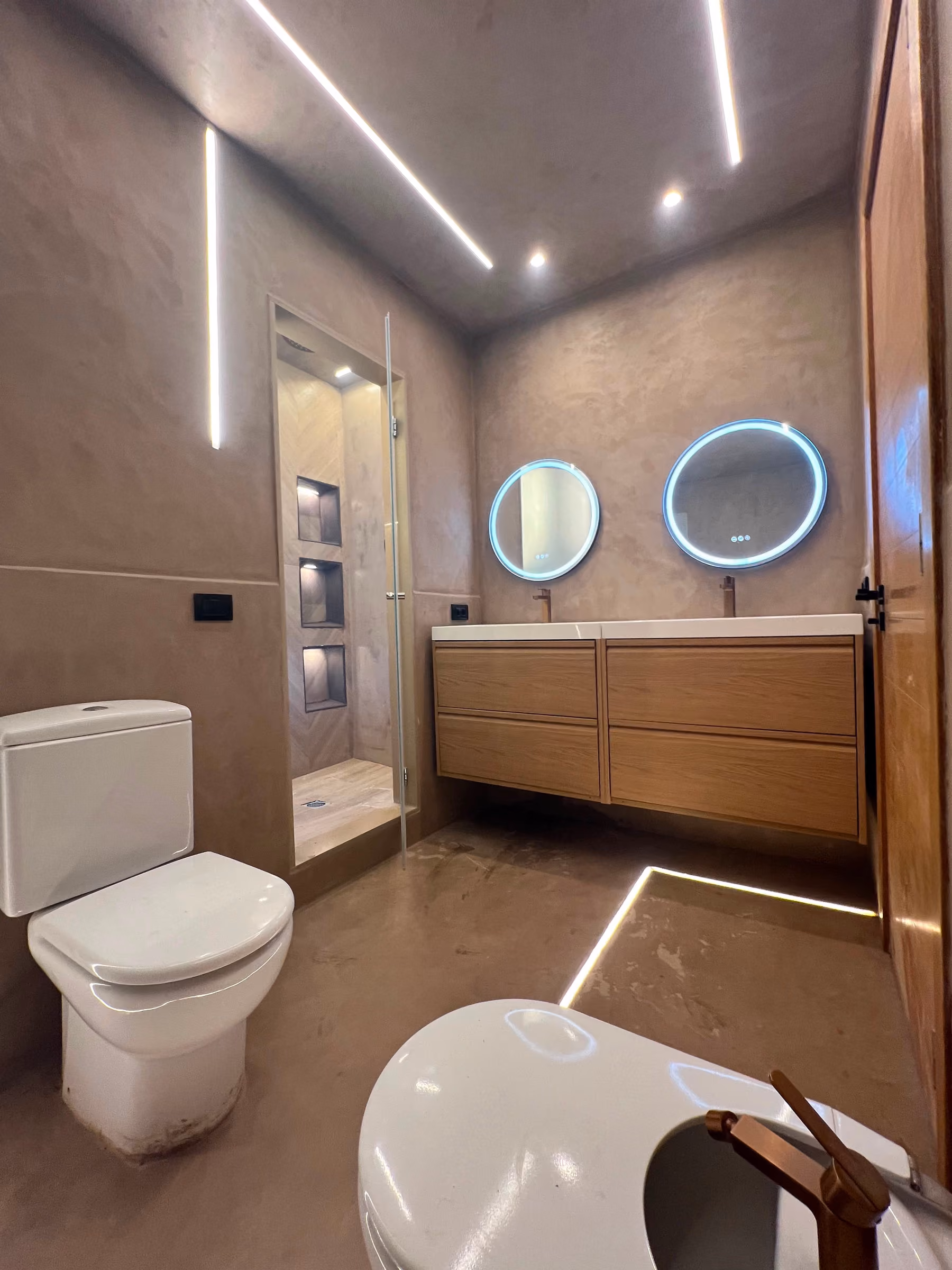 Baño en microcemento arena con lavabos gemelos, espejos circulares retroiluminados y ducha con nichos