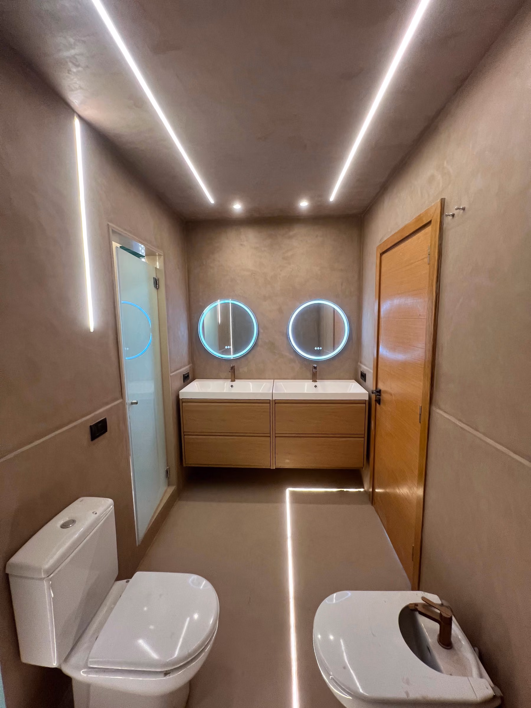 Vista general del baño en microcemento arena con doble lavabo y puerta de madera