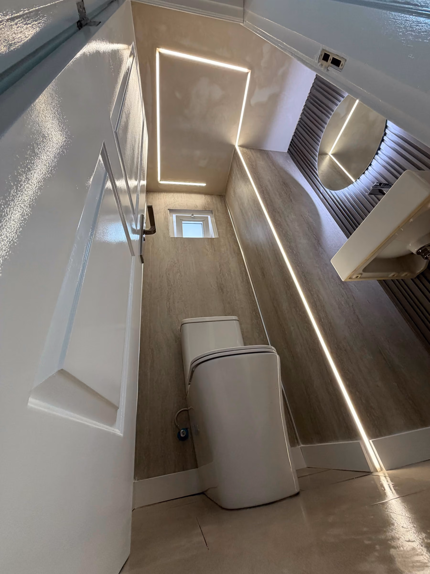 Aseo en travertino con inodoro, ventana y espejo circular integrado en el revestimiento