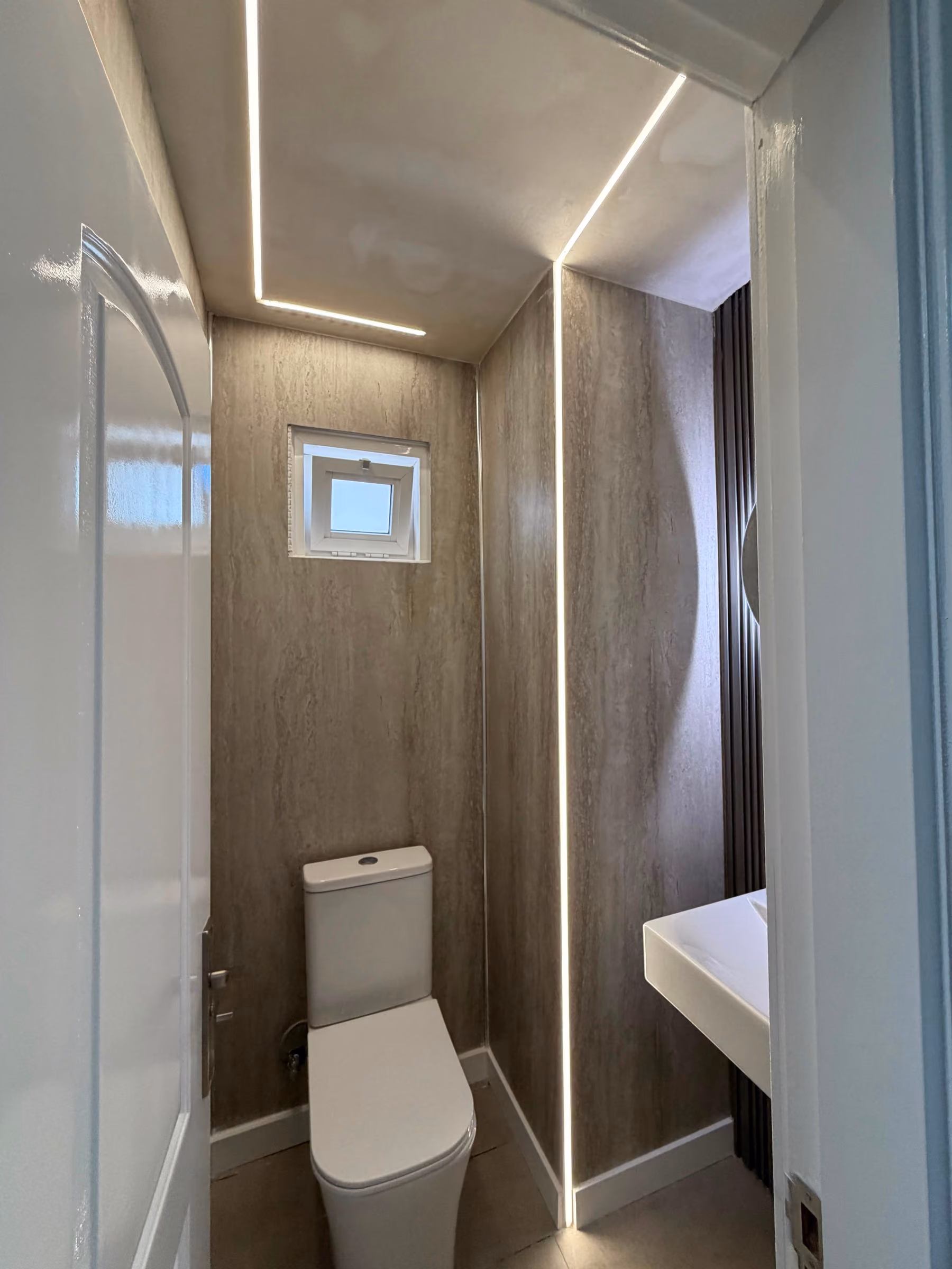 Aseo en travertino con inodoro, ventana y lavabo suspendido junto a panel oscuro acanalado