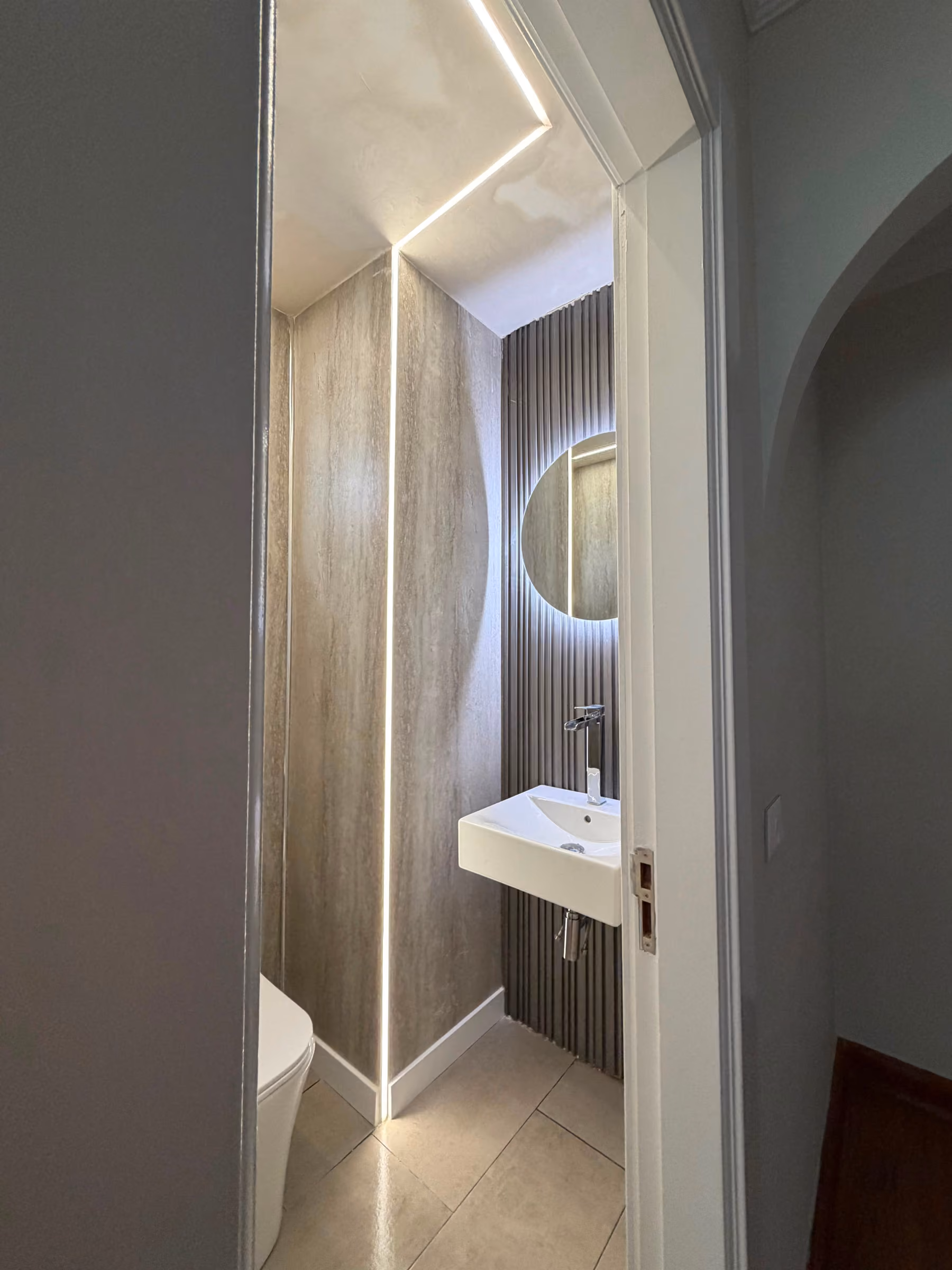 Lavabo suspendido bajo espejo circular en aseo de travertino con panel acanalado