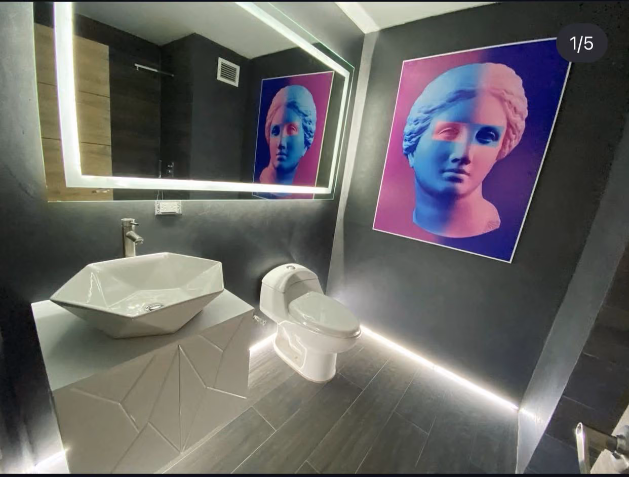 Vista general del baño con lavabo facetado, inodoro y arte gráfico enmarcado junto a espejo retroiluminado
